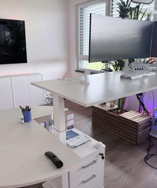Modern white home office with a curved desk, three mounted monitors, keyboard, and speakers; purple LED under the desk and a plant by the window.