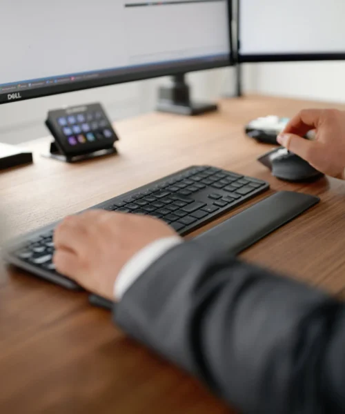 Person in a suit typing on a keyboard and using a mouse at a wooden desk with two computer monitors in view (office setup).